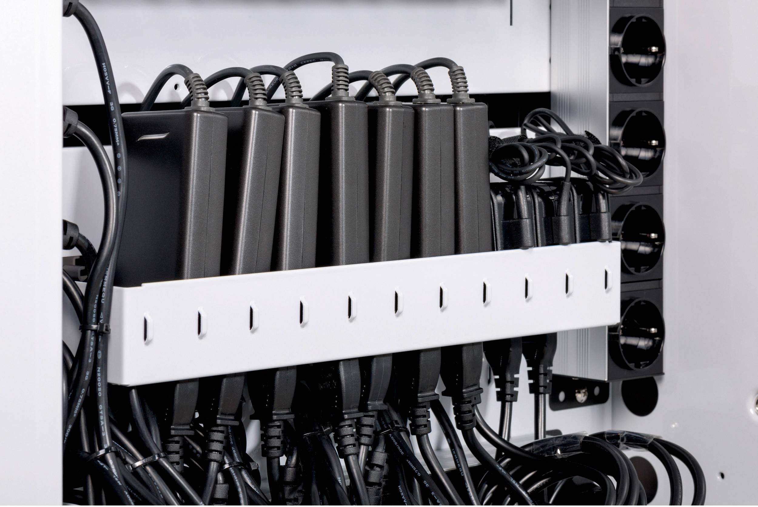 Mehrere schwarze Netzteile sind ordentlich in einer weißen Halterung in einem Schrank organisiert, umgeben von Kabeln und Steckdosenleisten.