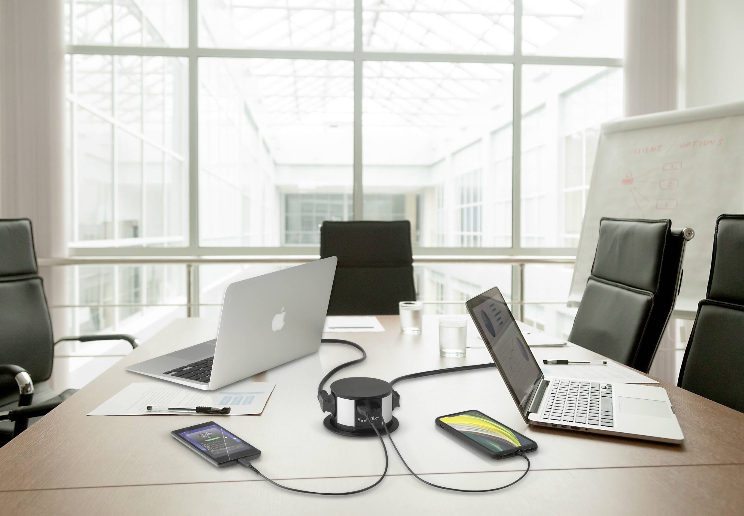 Ein moderner Konferenzraum mit zwei Laptops, Handys und einem zentralen Verbindungshub auf einem Tisch. Große Fenster im Hintergrund.