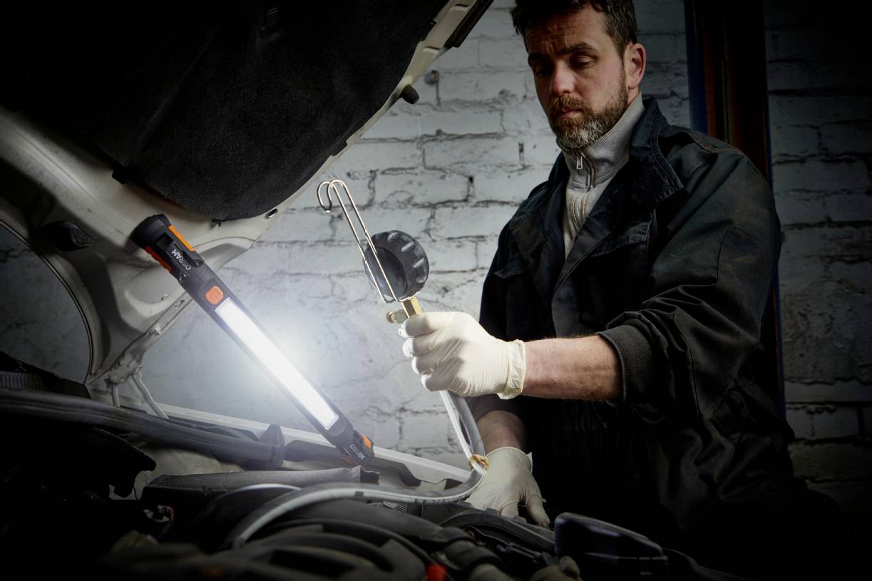 Mechaniker überprüft Motor eines Autos in einer Werkstatt. Er trägt Handschuhe und hält ein Arbeitslicht, das auf den Motor gerichtet ist.