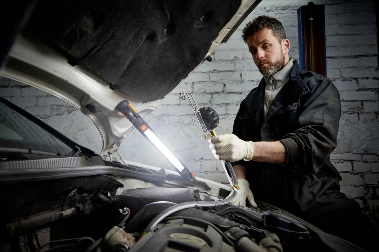 Mechaniker repariert Motor, steht an offenem Auto mit Arbeitslampe. Werkstatt-Umgebung, Ziegelwand im Hintergrund.