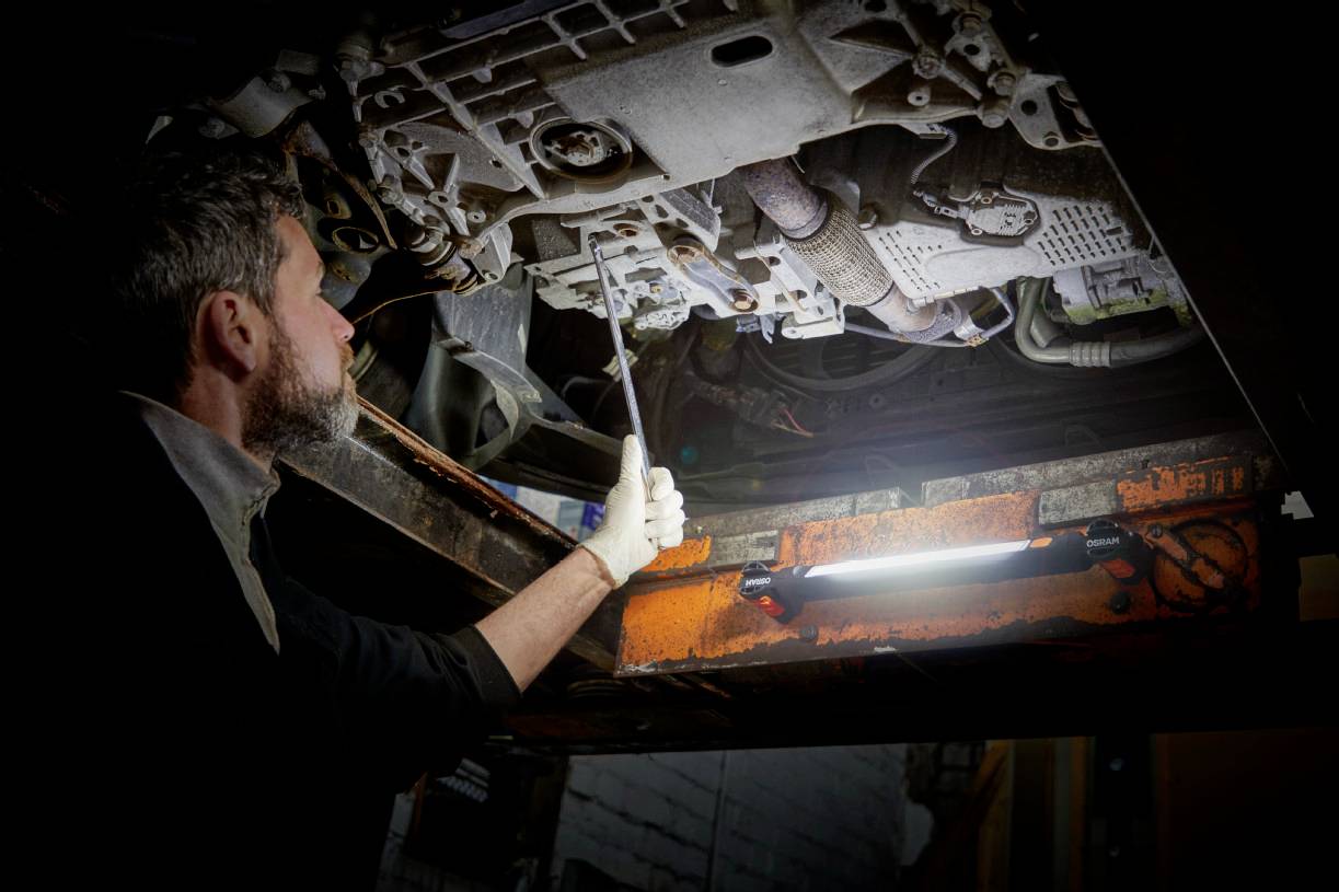 Ein Mechaniker inspiziert die Unterseite eines Fahrzeugs in einer Werkstatt. Er benutzt eine Leuchte für bessere Sichtbarkeit.