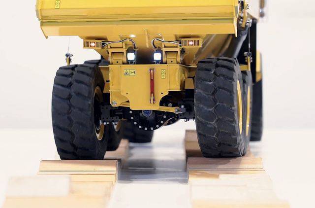 Großer gelber Muldenkipper fährt über hölzerne Platten. Die Aufnahme zeigt das Fahrzeug von hinten auf hellem Untergrund.