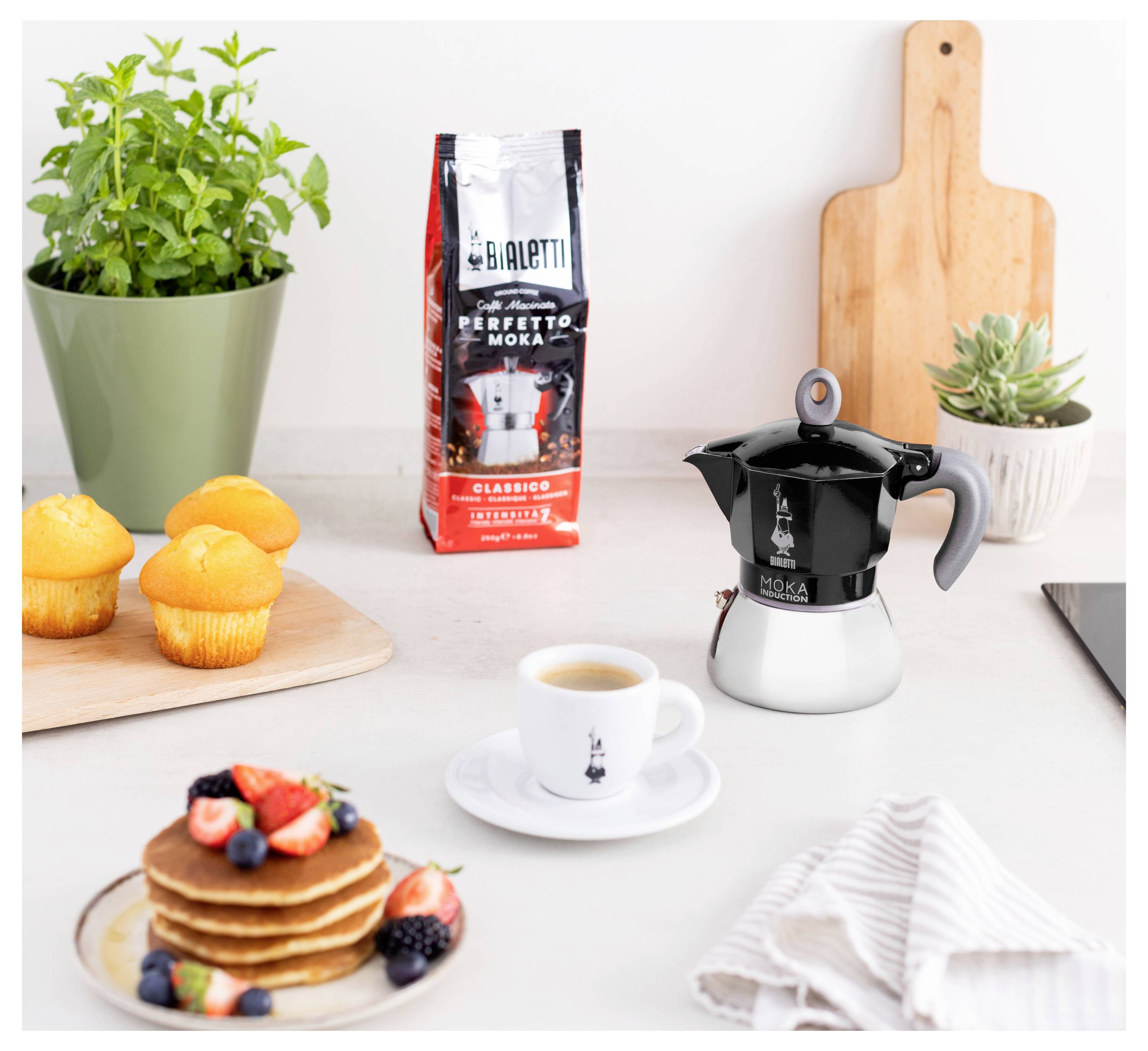 Eine Bialetti Mokka-Kanne und eine Kaffeetasse stehen auf einer Küchenarbeitsplatte, daneben Pfannkuchen, Muffins und eine Tüte 'Perfetto Moka' Kaffee.