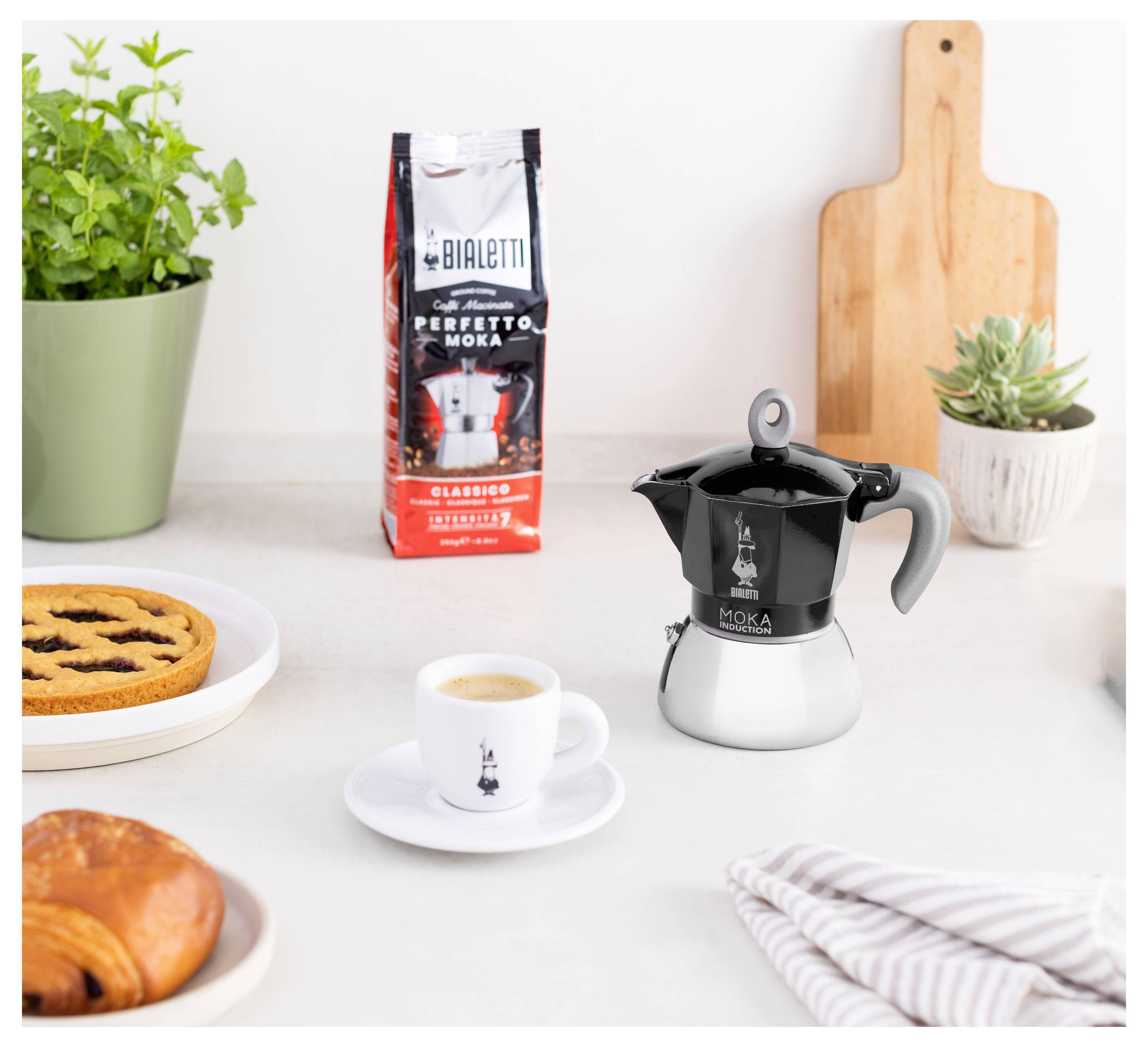 Eine Kaffeemaschine auf einer Küchenarbeitsplatte mit einer Tasse Espresso, einer Tüte Bialetti 'Perfetto Moka' Kaffee, einem Gebäckstück und Pflanzen in der Nähe.