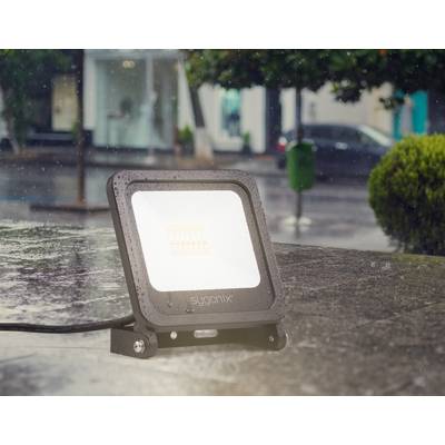 Eine schwarze LED-Flutlichtlampe steht im Regen auf einem Tisch im Freien. Im Hintergrund sind Bäume und eine regnerische Straße zu sehen.