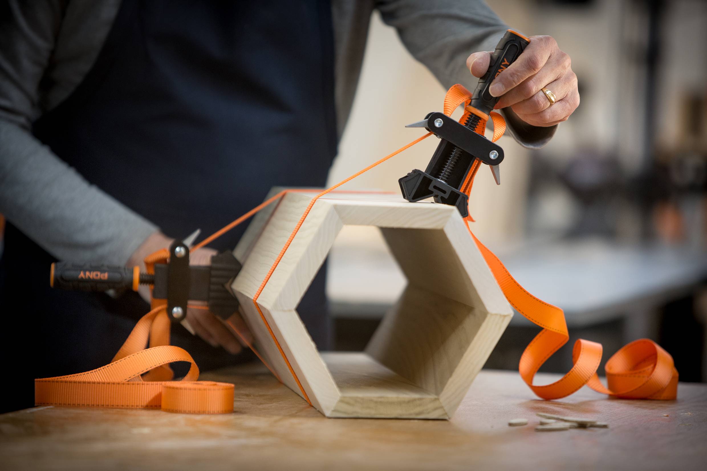 Eine Person spannt ein sechseckiges Holzstück mit orangenen Spanngurten fest, möglicherweise zur Stabilisierung beim Bau oder Arbeiten.