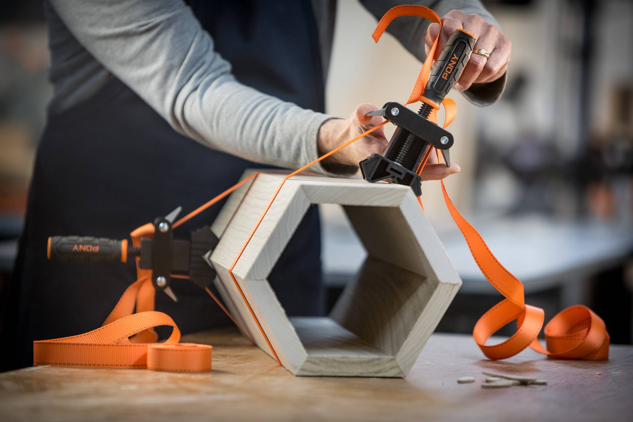 Eine Person befestigt ein orangefarbenes Band um ein sechseckiges Holzobjekt mit Klemmen auf einem Tisch, möglicherweise beim Heimwerken.