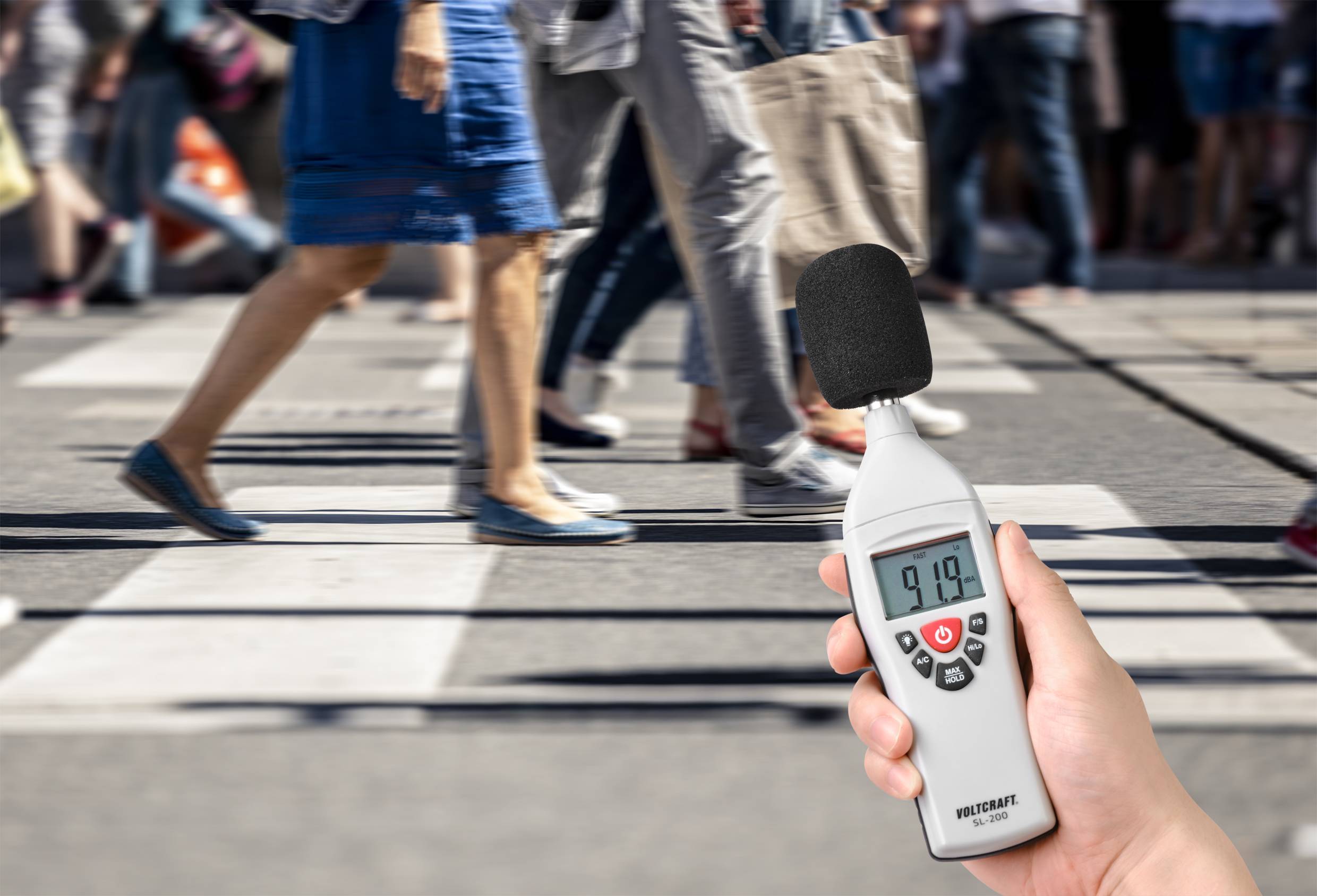 Eine Person hält ein Schallpegelmessgerät, das 91,9 Dezibel anzeigt, während im Hintergrund Menschen eine belebte Straße überqueren.