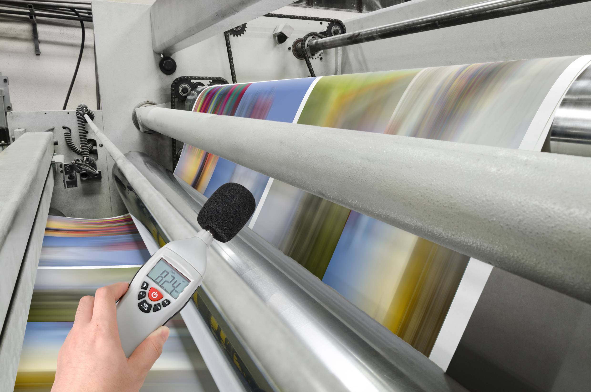 Druckmaschine, die farbige Seiten produziert. Ein Dezibelmeter misst den Geräuschpegel bei 82,1 dB in der Nähe der Maschine.