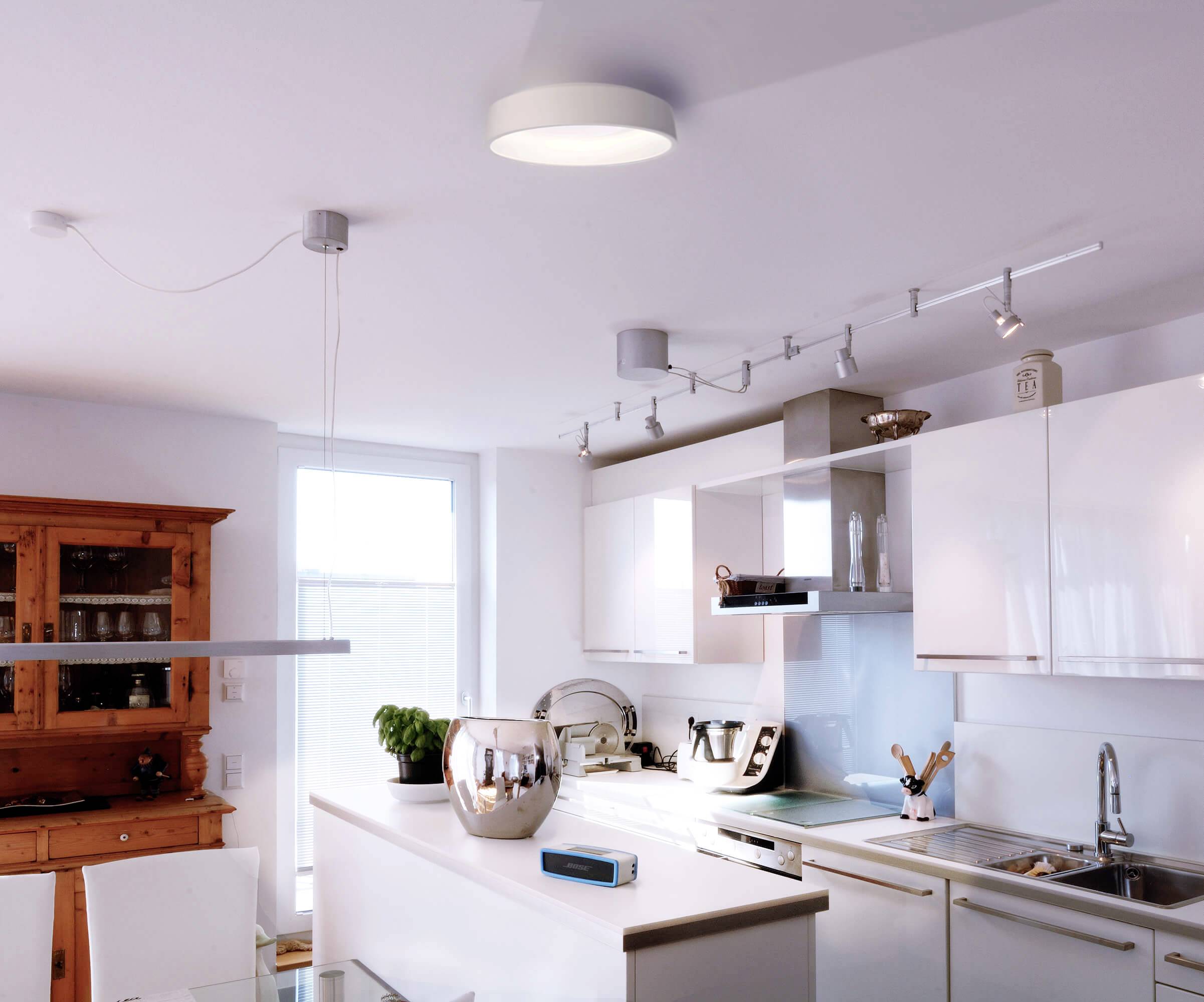 Moderne Küche mit weißer Einrichtung. LED-Leuchten an der Decke, Küchenutensilien auf der Arbeitsfläche. Helles Tageslicht.