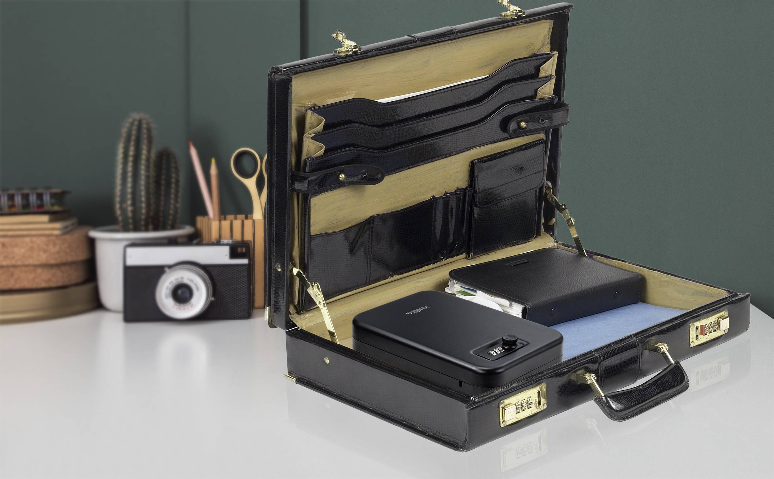 Ein geöffneter schwarzer Aktenkoffer auf einem Schreibtisch. Darin befinden sich Ordner und ein elektronisches Gerät. Im Hintergrund sind Schreibutensilien und ein Fotoapparat.