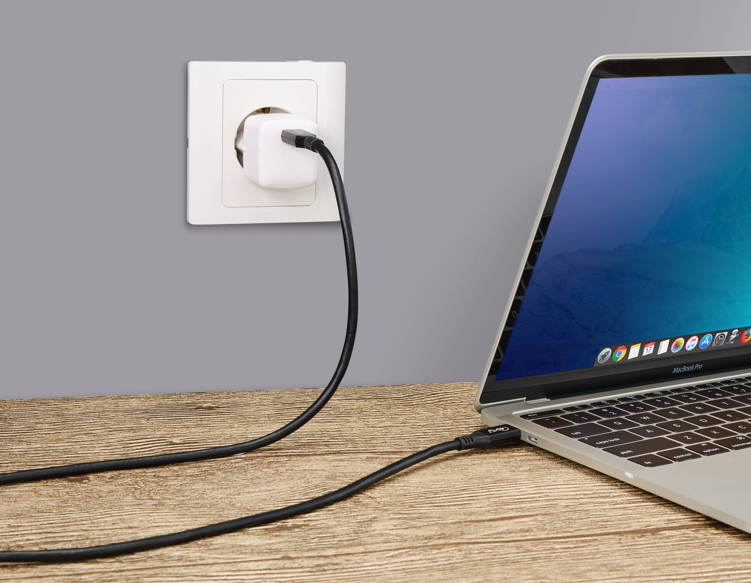 Ein Laptop wird über ein Ladekabel, das an einer Wandsteckdose angeschlossen ist, aufgeladen. Der Bildschirm zeigt den Desktop-Hintergrund.