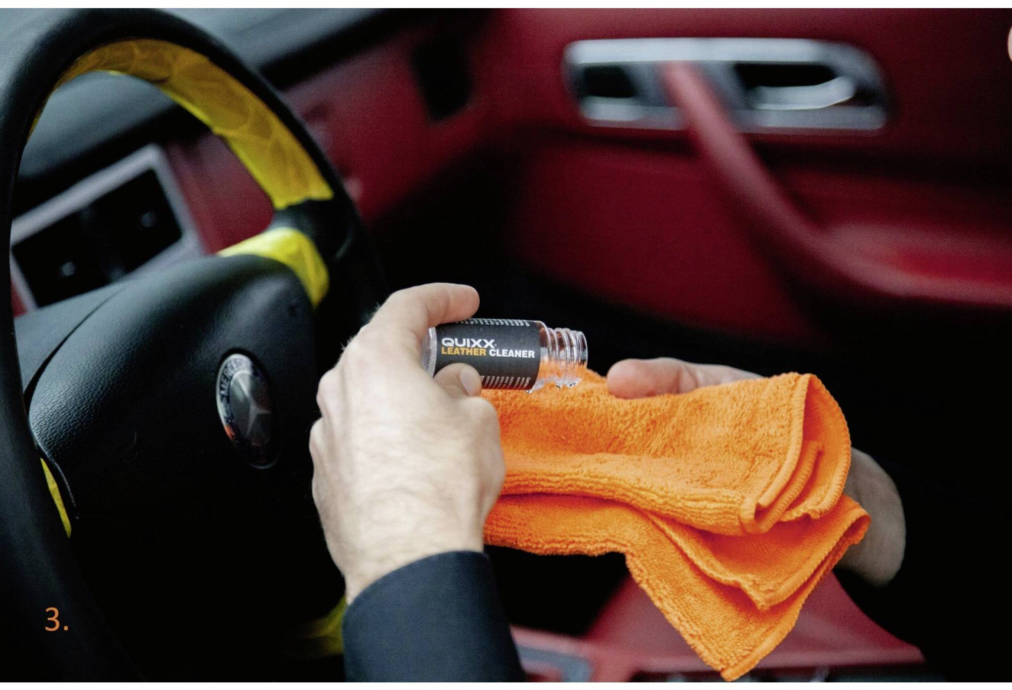 Person wischt Armaturenbrett eines Autos mit einem orangefarbenen Tuch und einer Reinigungsflüssigkeit in einer Flasche ab.