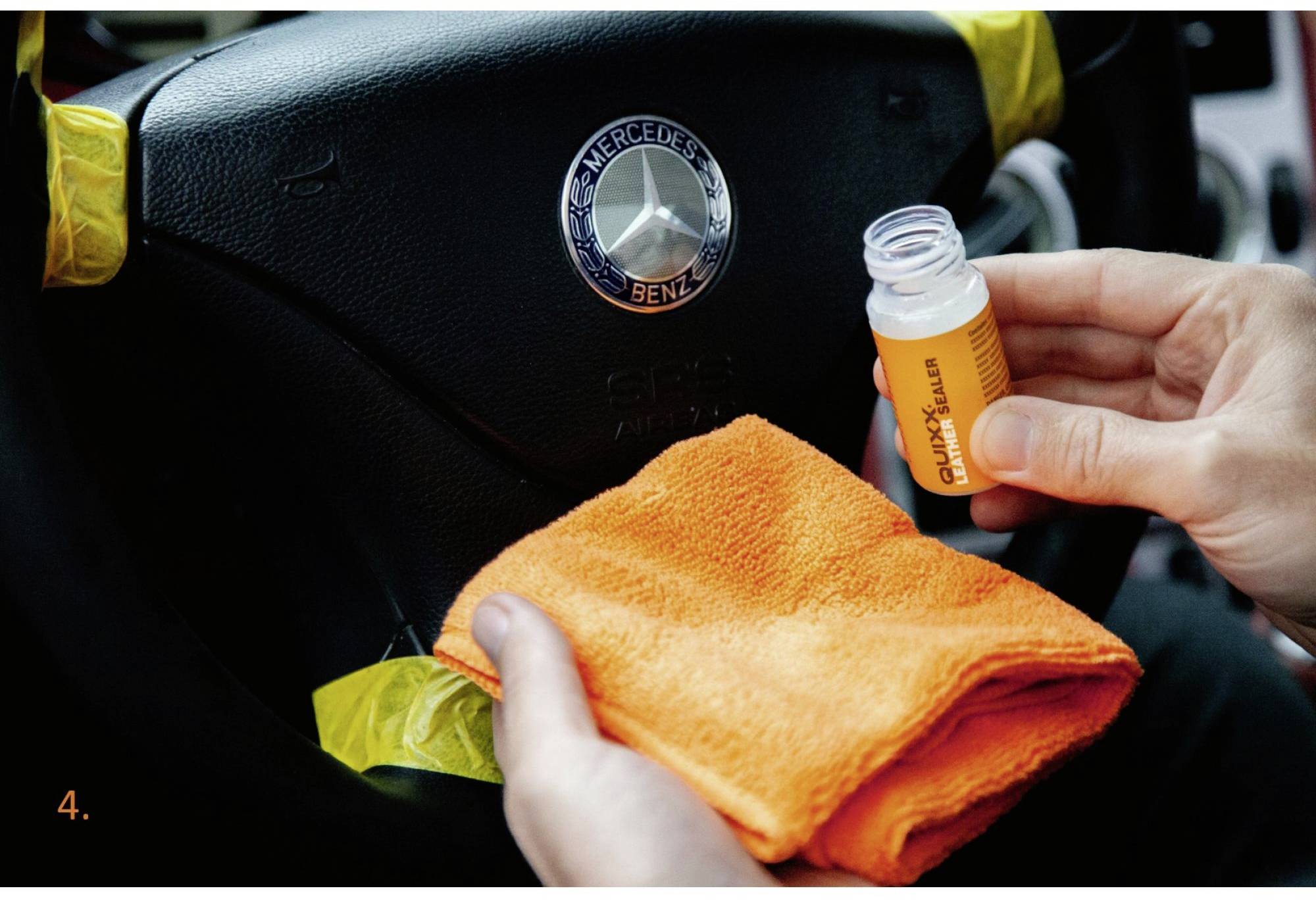 Person reinigt ein Lenkrad mit einem orangefarbenen Tuch und einer kleinen Flasche Reiniger. Mercedes-Logo sichtbar auf dem Lenkrad.