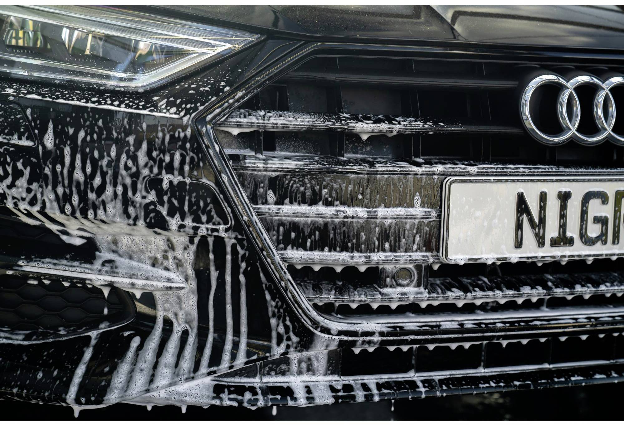 In Nahaufnahme zeigt das Bild einen schwarzen Audi mit Schaum beim Autowaschen, fokussiert auf den Kühlergrill und das Autokennzeichen.