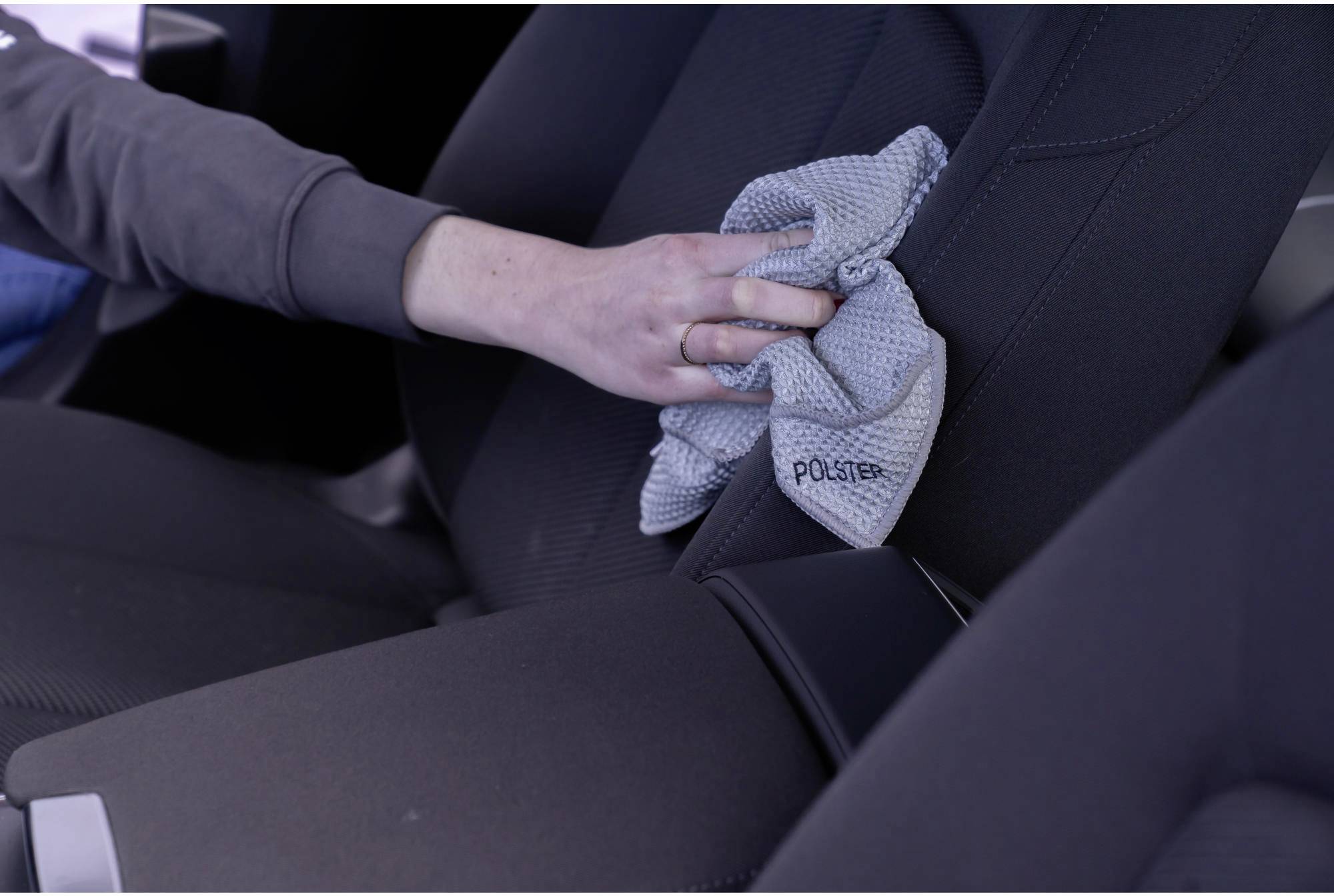 Eine Person reinigt mit einem grauen Tuch den Autositz eines Fahrzeugs. Sie trägt ein langärmliges Oberteil. Der Fokus liegt auf der Reinigungshandlung.