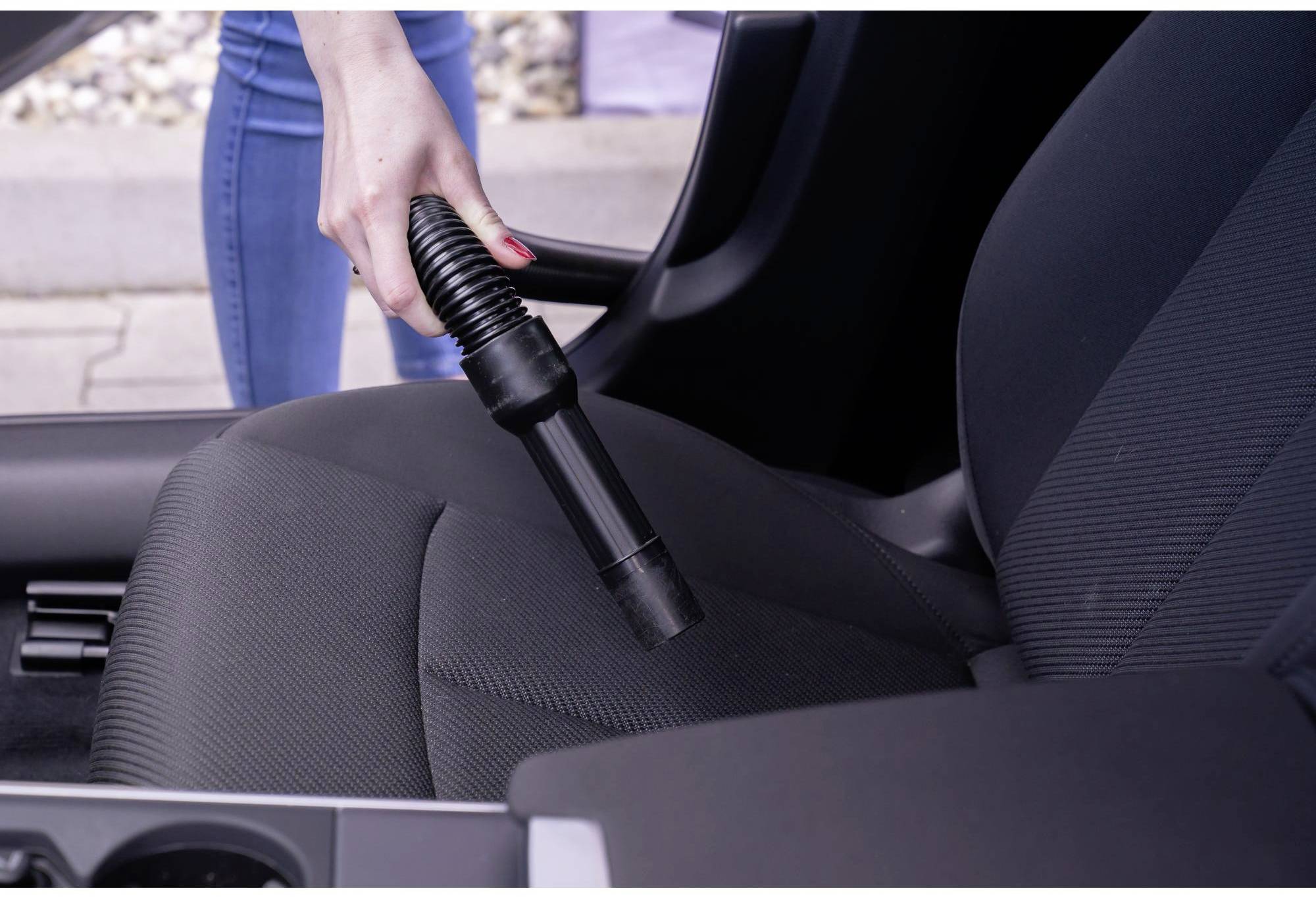 Eine Person saugt den Sitz eines Autos mit einem Staubsauger. Der Fokus liegt auf der Hand, die den Staubsaugerschlauch hält.