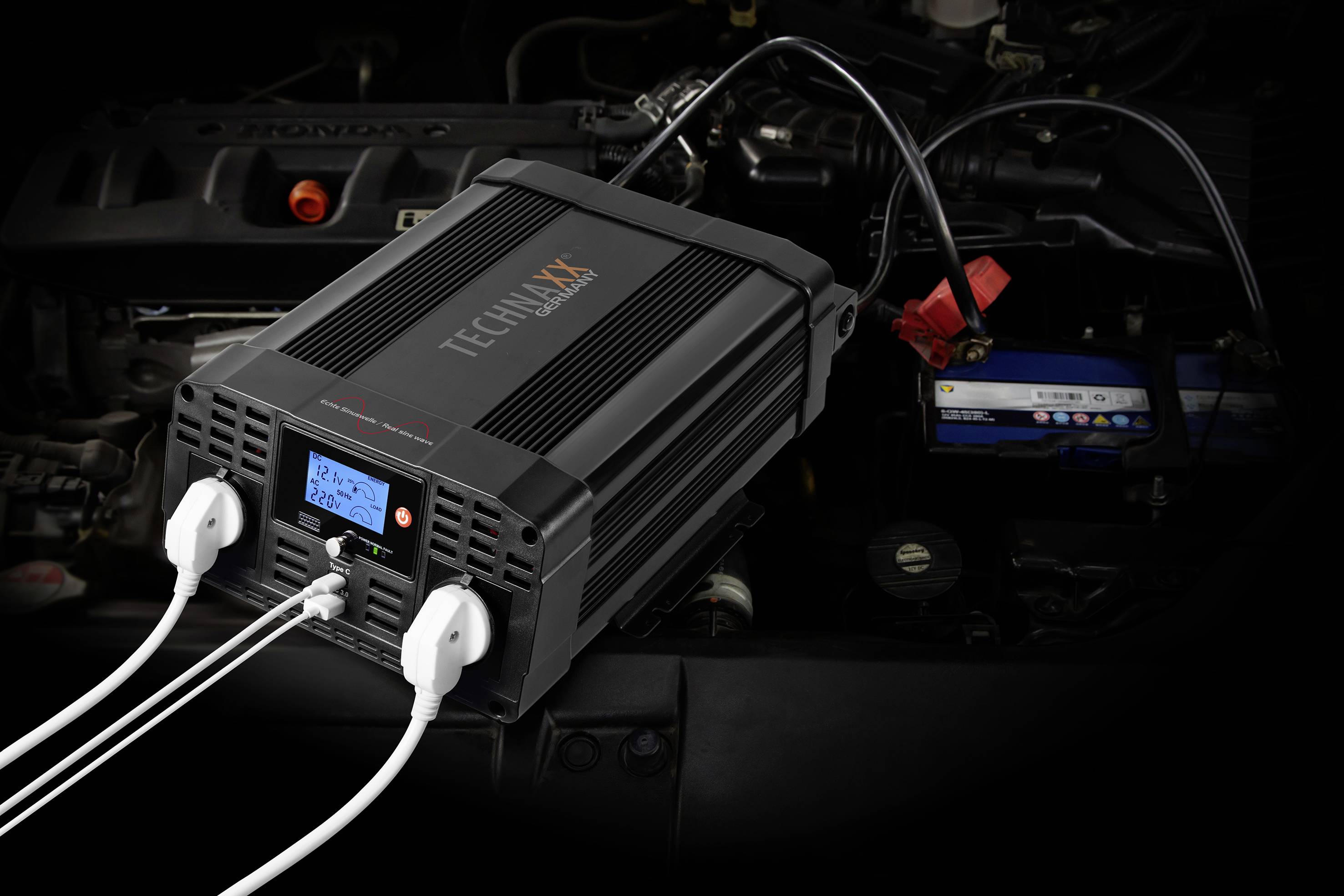 Ein Auto-Batterieladegerät von Technaxx zeigt die Anzeige mit zwei angeschlossenen Kabeln, die an eine Autobatterie im Motorraum führen.