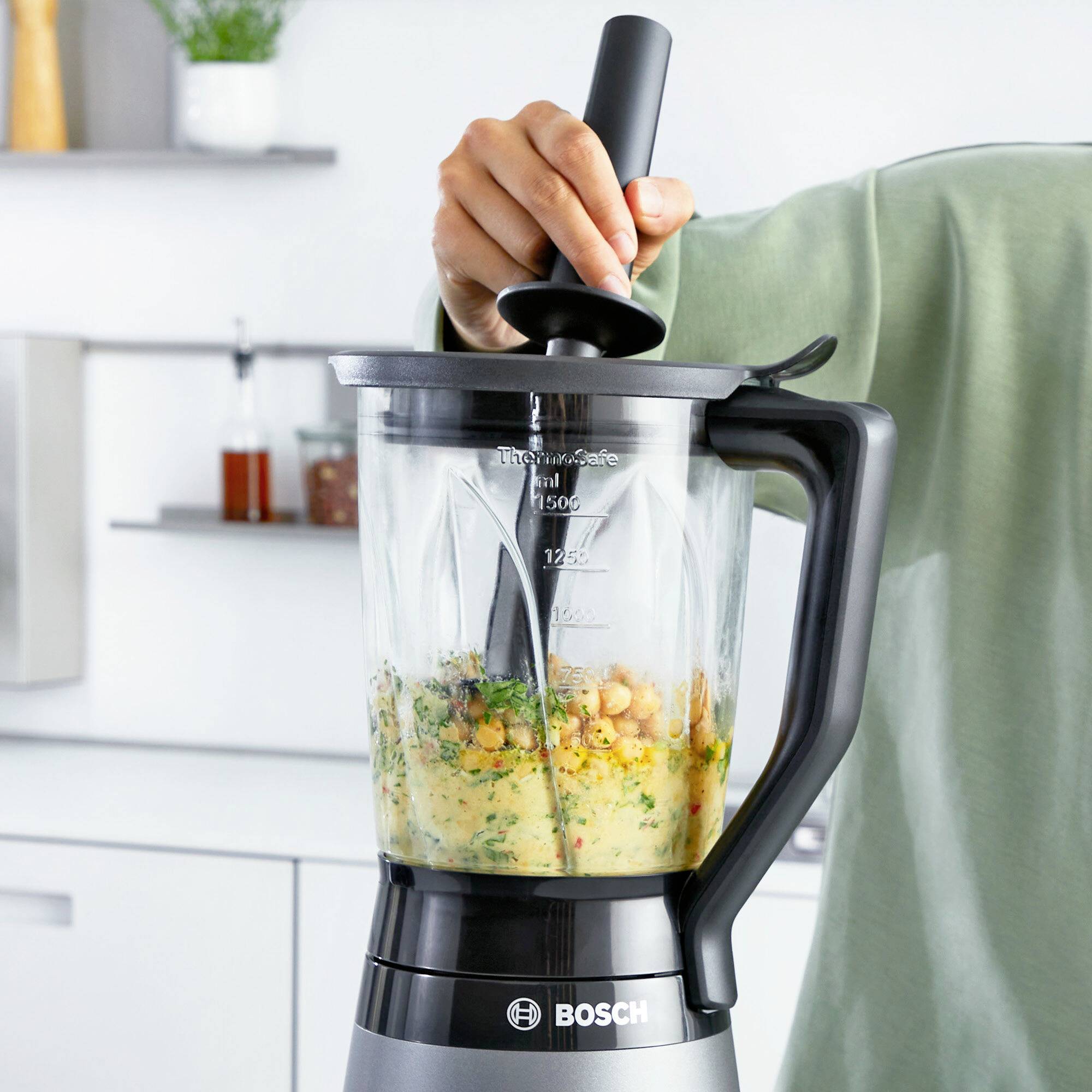 Eine Hand bedient einen Bosch-Standmixer, der Gemüsemix zerkleinert. Die Küche im Hintergrund ist modern und hell gestaltet.