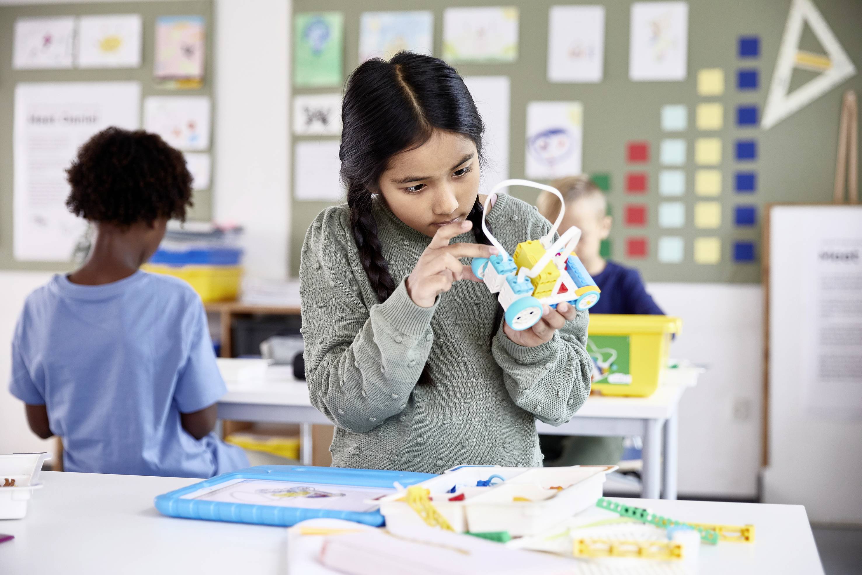 Ein Mädchen im Klassenzimmer untersucht ein kleines Modellauto aus bunten Bausteinen. Im Hintergrund arbeiten weitere Kinder an Projekten.