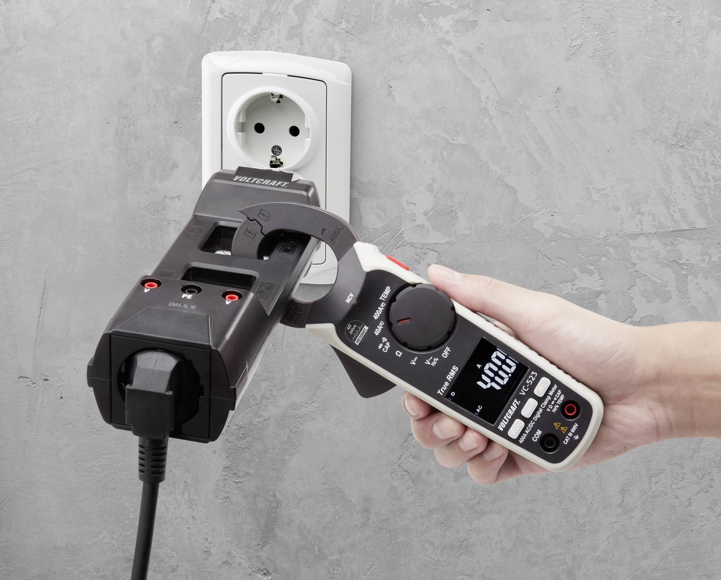 Eine Hand hält ein digitales Multimeter und misst Stromstärke an einem Steckdosenadapter, der in eine Wandsteckdose eingesteckt ist.