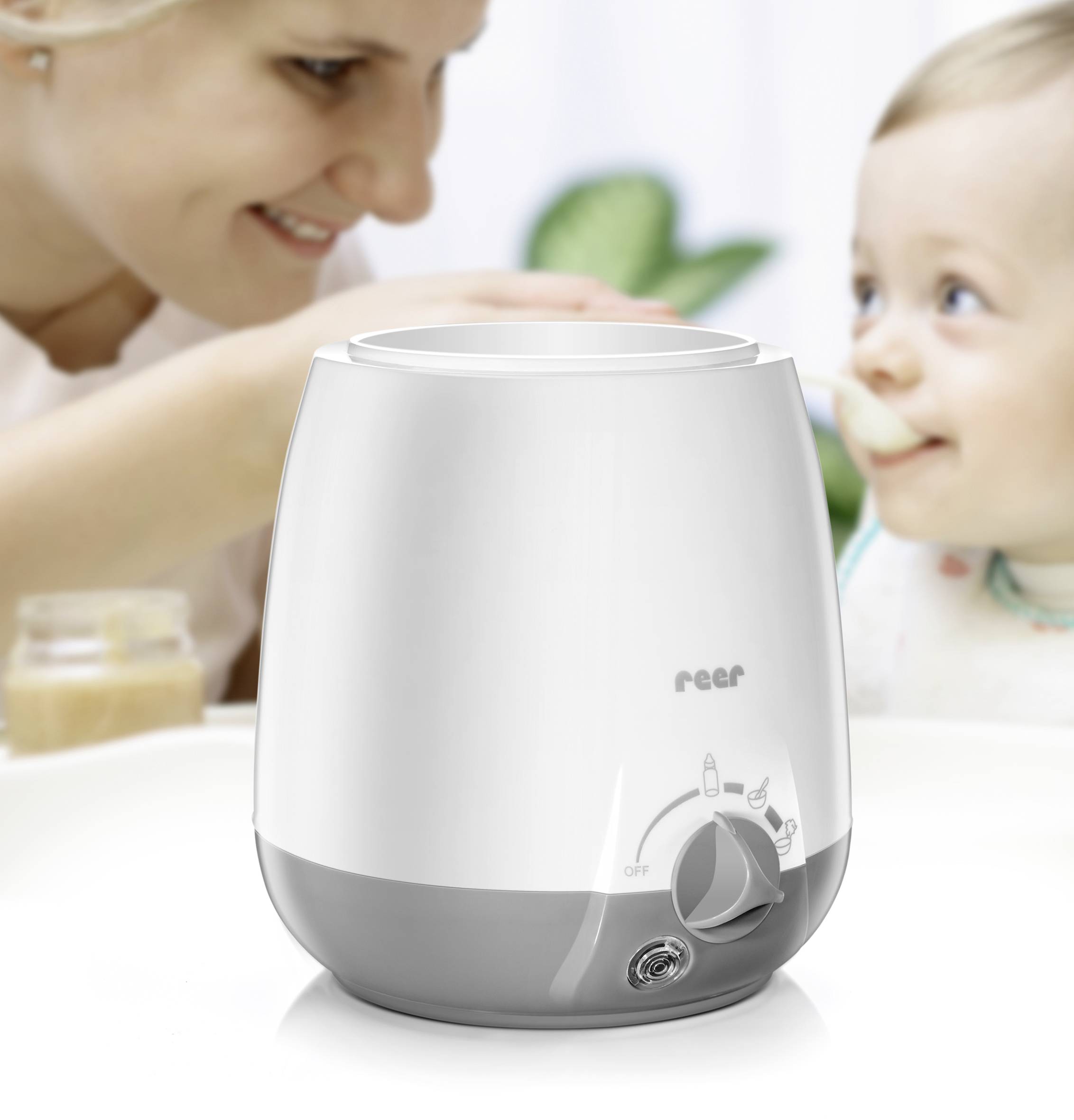 Eine Frau füttert ein Kleinkind im Hintergrund; im Vordergrund steht ein Babykostwärmer auf einem Tisch.