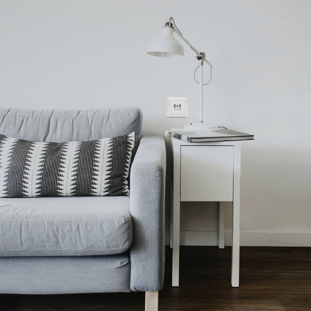 Graues Sofa mit gemustertem Kissen neben einem weißen Nachttisch mit Lampe. Ruhiges, minimalistisches Wohnzimmer.