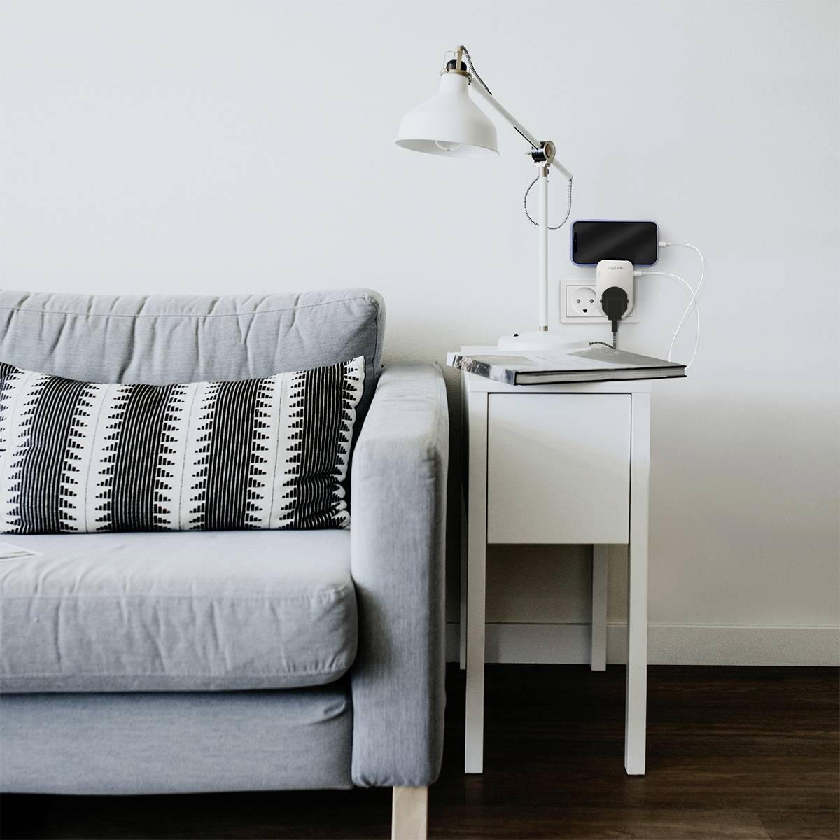 Ein graues Sofa mit zwei gemusterten Kissen steht neben einem weißen Tisch, auf dem eine Lampe und ein aufladendes Smartphone liegen.