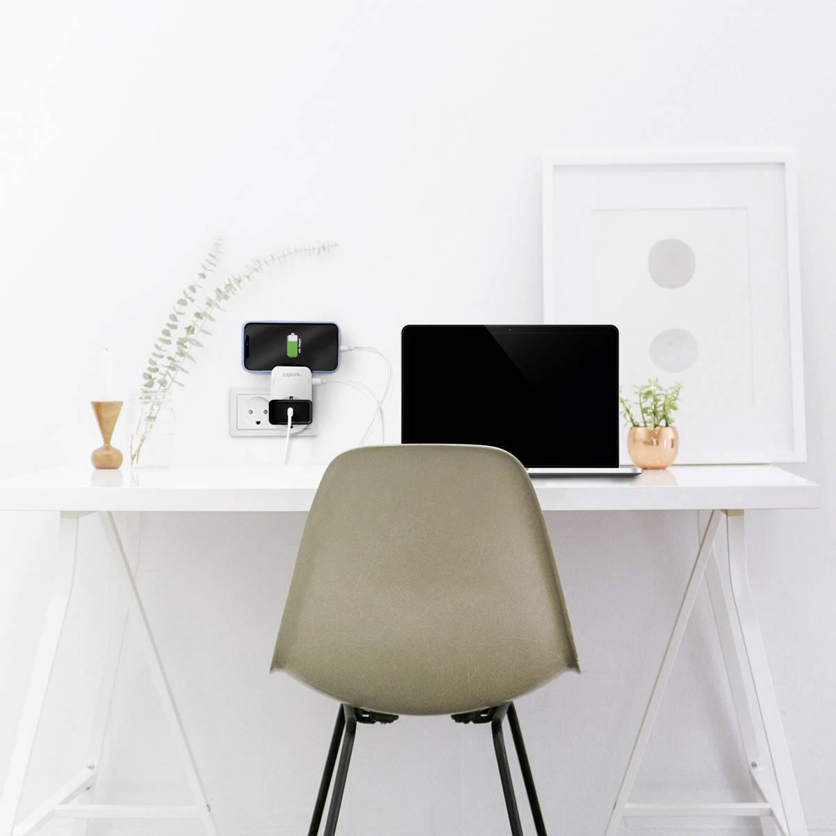 Ein moderner Schreibtisch mit einem Laptop, einer Wandsteckdose mit eingestecktem Handy, Pflanzenvasen und minimalistischen Dekorationen.