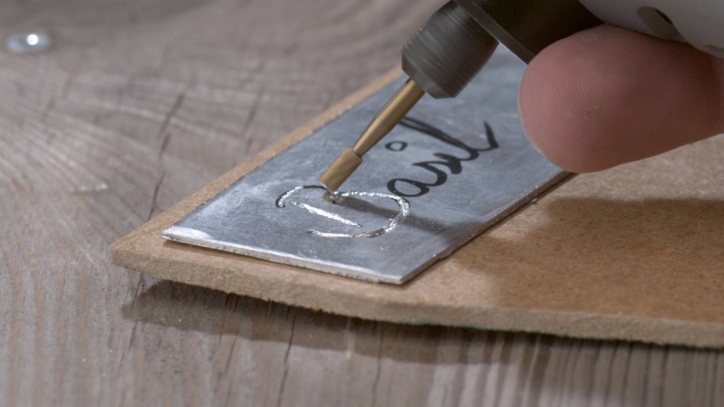 Eine Hand halt einen Gravierstift und graviert das Wort 'Basil' auf eine Metallplatte, die auf einem Holztisch liegt.