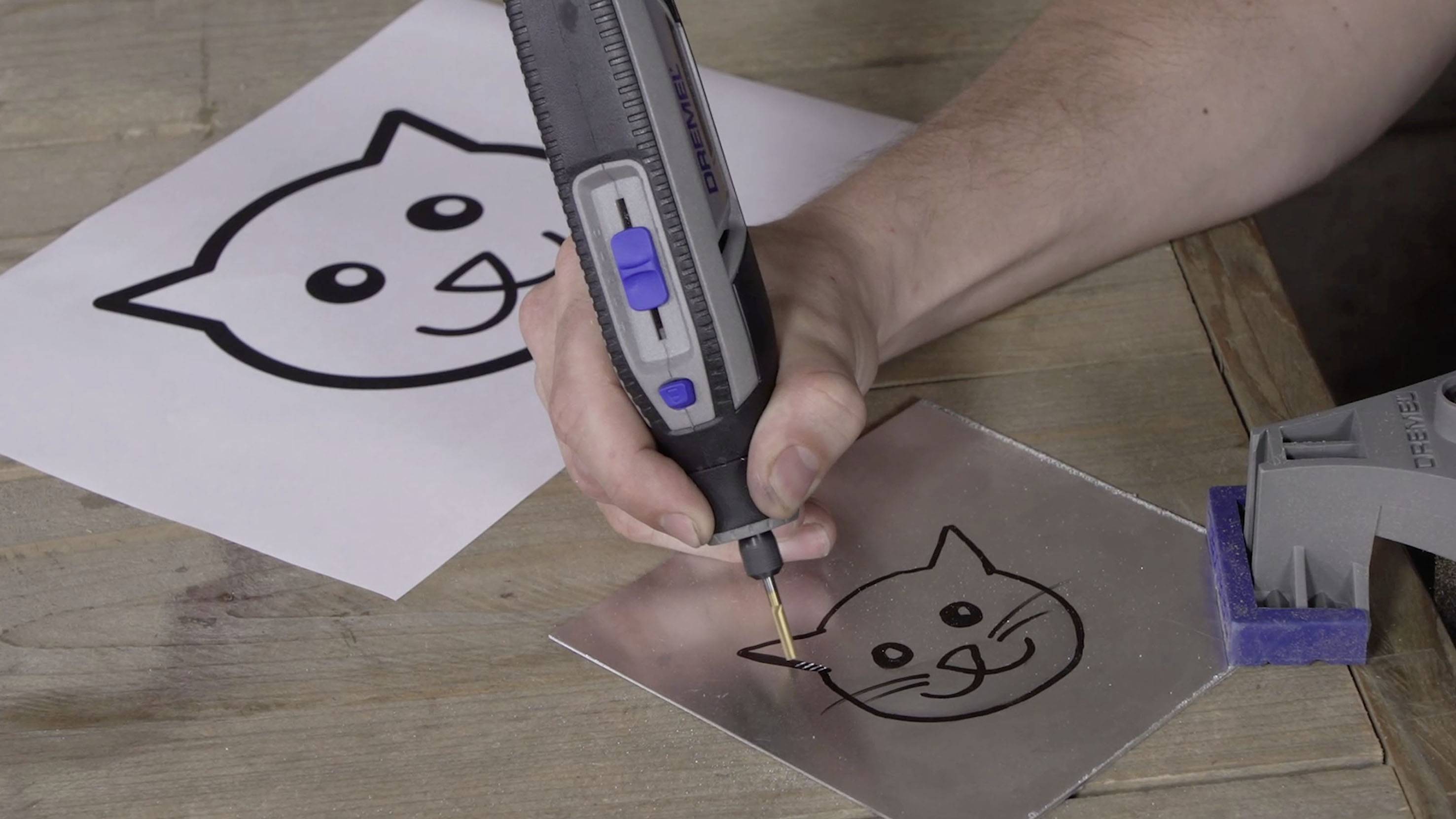 Eine Person zeichnet mit einem elektrischen Werkzeug das Bild eines Katzenkopfes auf eine Metallplatte, neben einer Papiervorlage.
