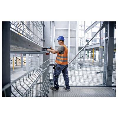 Ein Bauarbeiter mit blauem Helm und orangefarbener Warnweste bedient ein Elektrowerkzeug an einem Metallrahmen auf einer Baustelle.