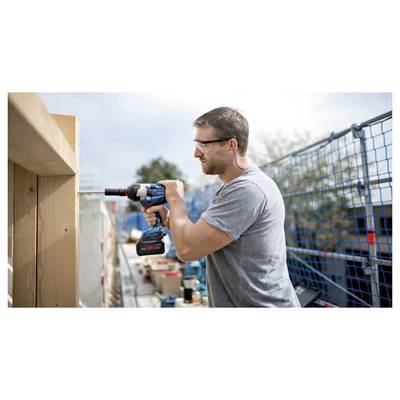 Ein Mann mit Sicherheitsbrille verwendet einen Akkuschrauber an einem Holzbalken auf einer Baustelle, umgeben von Sicherheitsabsperrungen und Bauausrüstung.