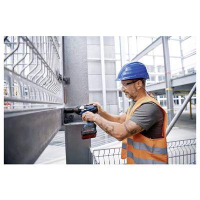 Ein Bauarbeiter mit blauem Helm und orangefarbener Warnweste verwendet ein Elektrowerkzeug, um einen Metallträger auf einer Baustelle zu befestigen.