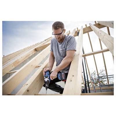 Eine Person mit Sicherheitsbrille bedient eine Bohrmaschine an Holzbalken eines sich im Bau befindlichen Daches vor einer teilweise bewölkten Himmelkulisse.