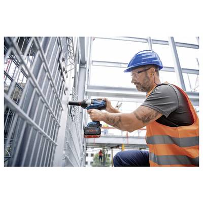 Ein Bauarbeiter mit blauem Schutzhelm und orangefarbener Sicherheitsweste bedient einen Bohrer an einer Metallkonstruktion auf einer Baustelle.