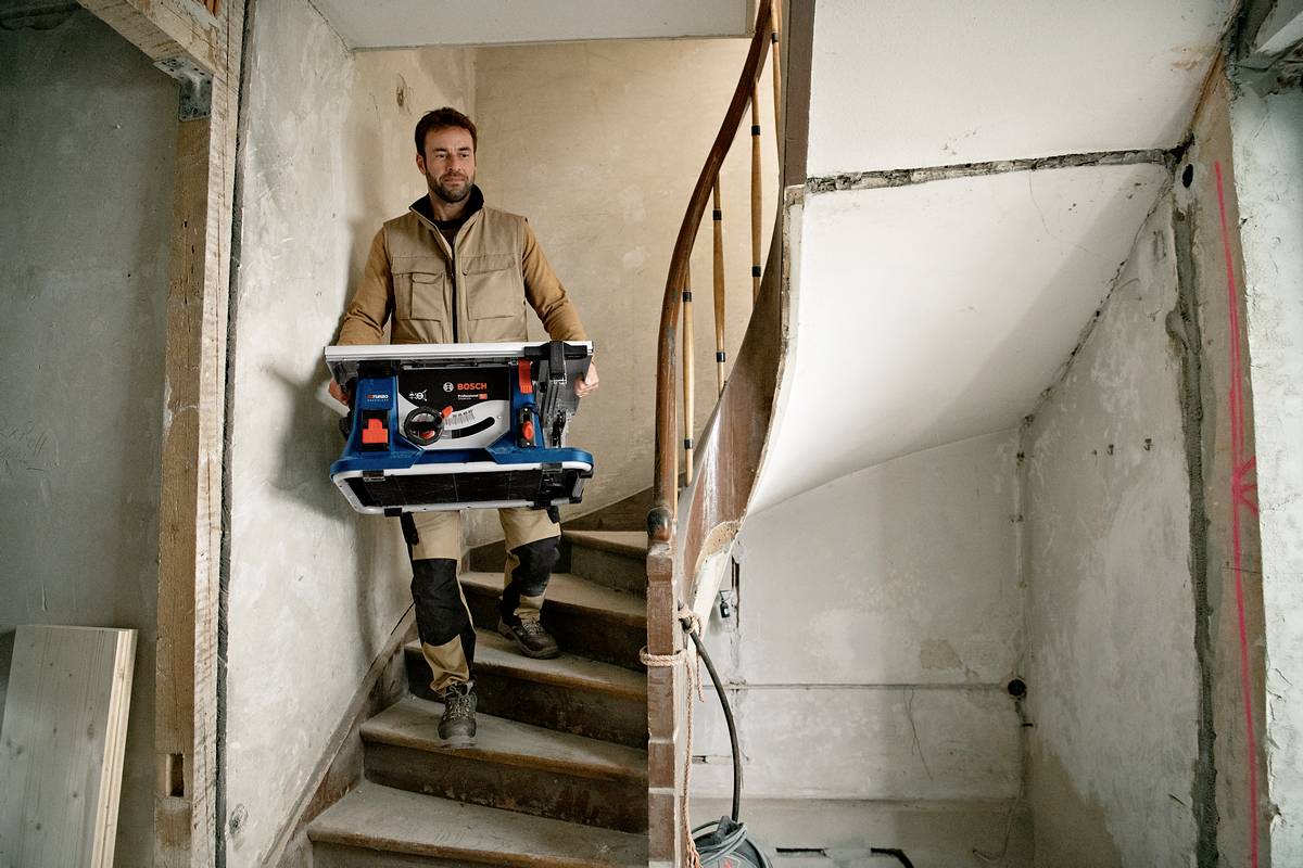 Ein Mann trägt eine Tischkreissäge die Treppe in einem unfertigen Gebäude hinauf. Er hält die Säge mit beiden Händen fest.