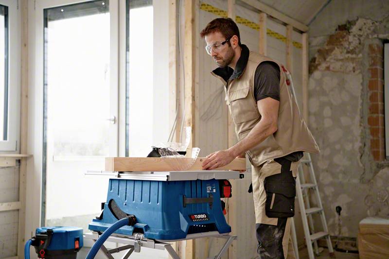 Ein Mann in Arbeitskleidung arbeitet an einem Tischkreissäge in einem Rohbau. Im Hintergrund ist ein Fenster und eine Leiter zu sehen.