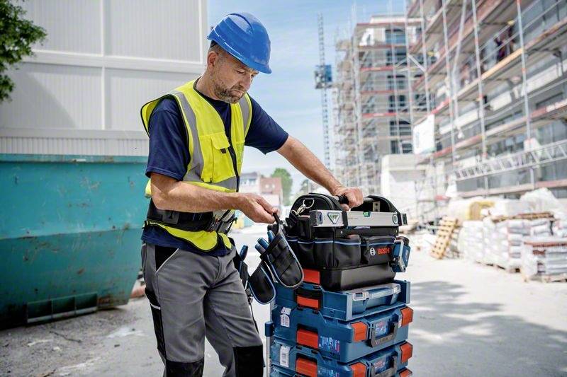 Ein Bauarbeiter mit Helm und Sicherheitsweste zieht eine Werkzeugbox auf einem Stapel von Kisten auf einer Baustelle. Im Hintergrund sind Baugerüste sichtbar.