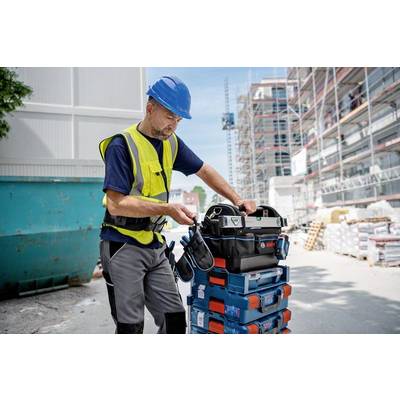 Ein Bauarbeiter mit Helm und Sicherheitsweste zieht eine Werkzeugbox auf einem Stapel von Kisten auf einer Baustelle. Im Hintergrund sind Baugerüste sichtbar.