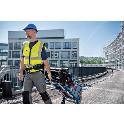 Ein Bauarbeiter trägt eine Sicherheitsweste und einen Helm. Er zieht eine Werkzeugtasche auf einem Rollwagen über einen Gehweg vor modernen Bürogebäuden.