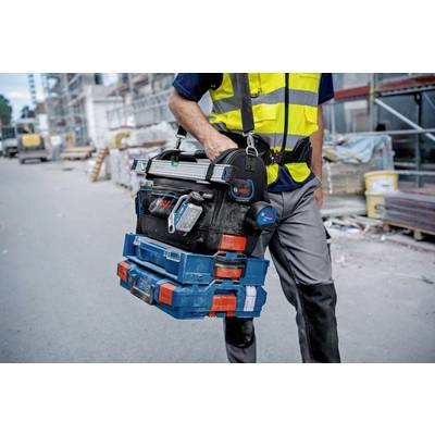 Arbeiter in Schutzweste, trägt Werkzeugkasten auf Baustelle. Hintergrund zeigt Baugerüst und Baumaterialstapel.