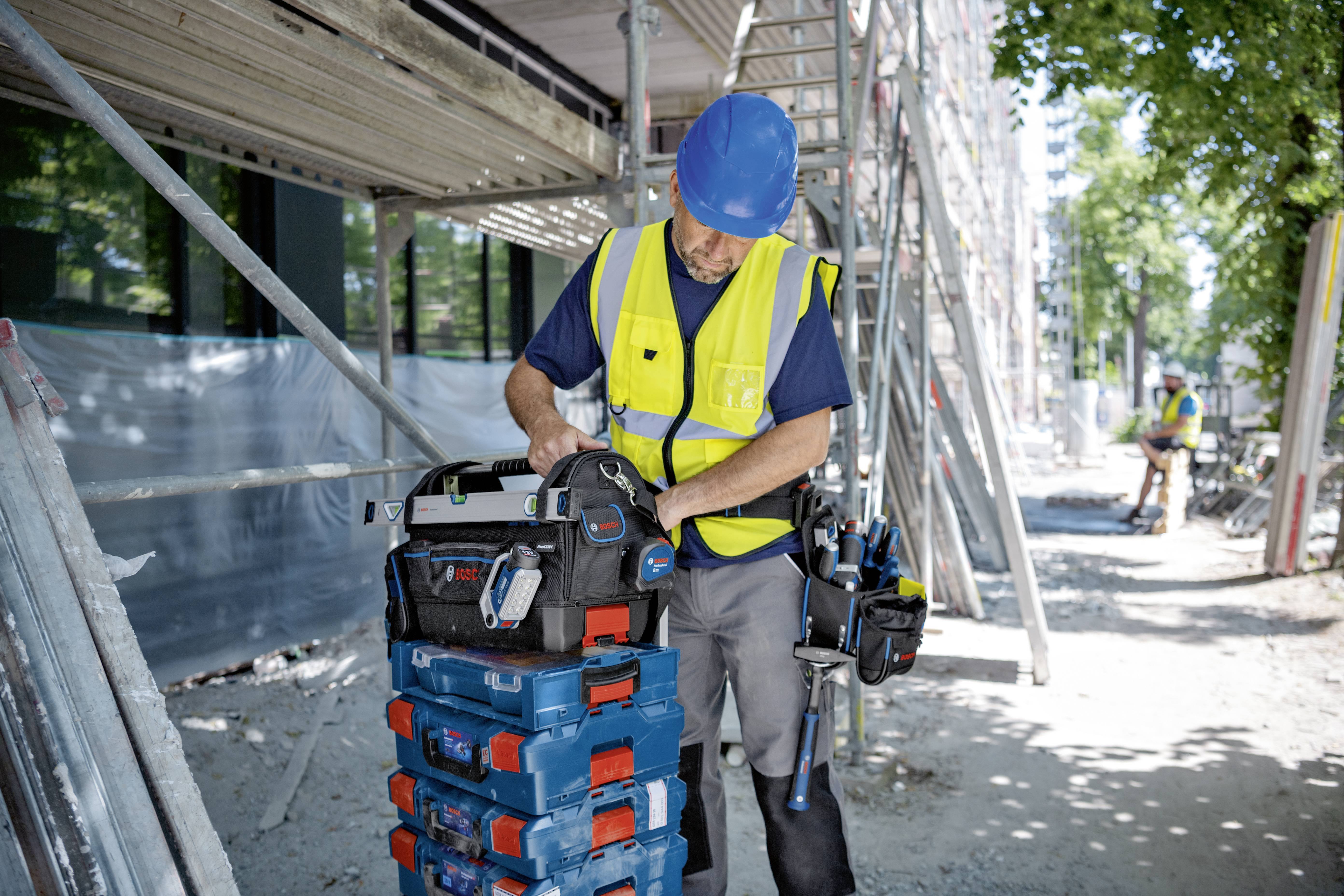 Ein Bauarbeiter mit blauem Helm und gelber Warnweste sortiert Werkzeugkoffer auf einer Baustelle vor einem Gerüst. Im Hintergrund ein weiterer Arbeiter.