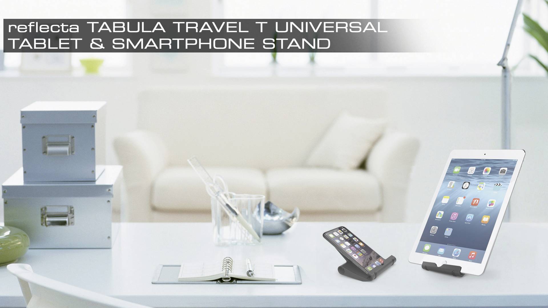 Auf einem Schreibtisch stehen ein Smartphone und ein Tablet auf Ständern. Im Hintergrund sind ein Sofa und Kisten zu sehen.