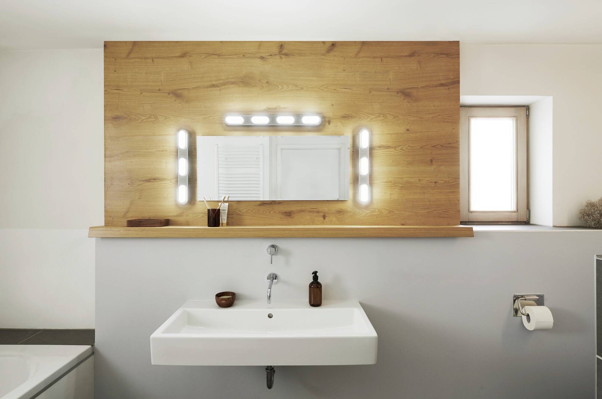 Ein modernes Badezimmer mit weißem Waschbecken, Holzwandverkleidung und beleuchtetem Spiegel. Links befinden sich Accessoires, rechts ein Fenster.