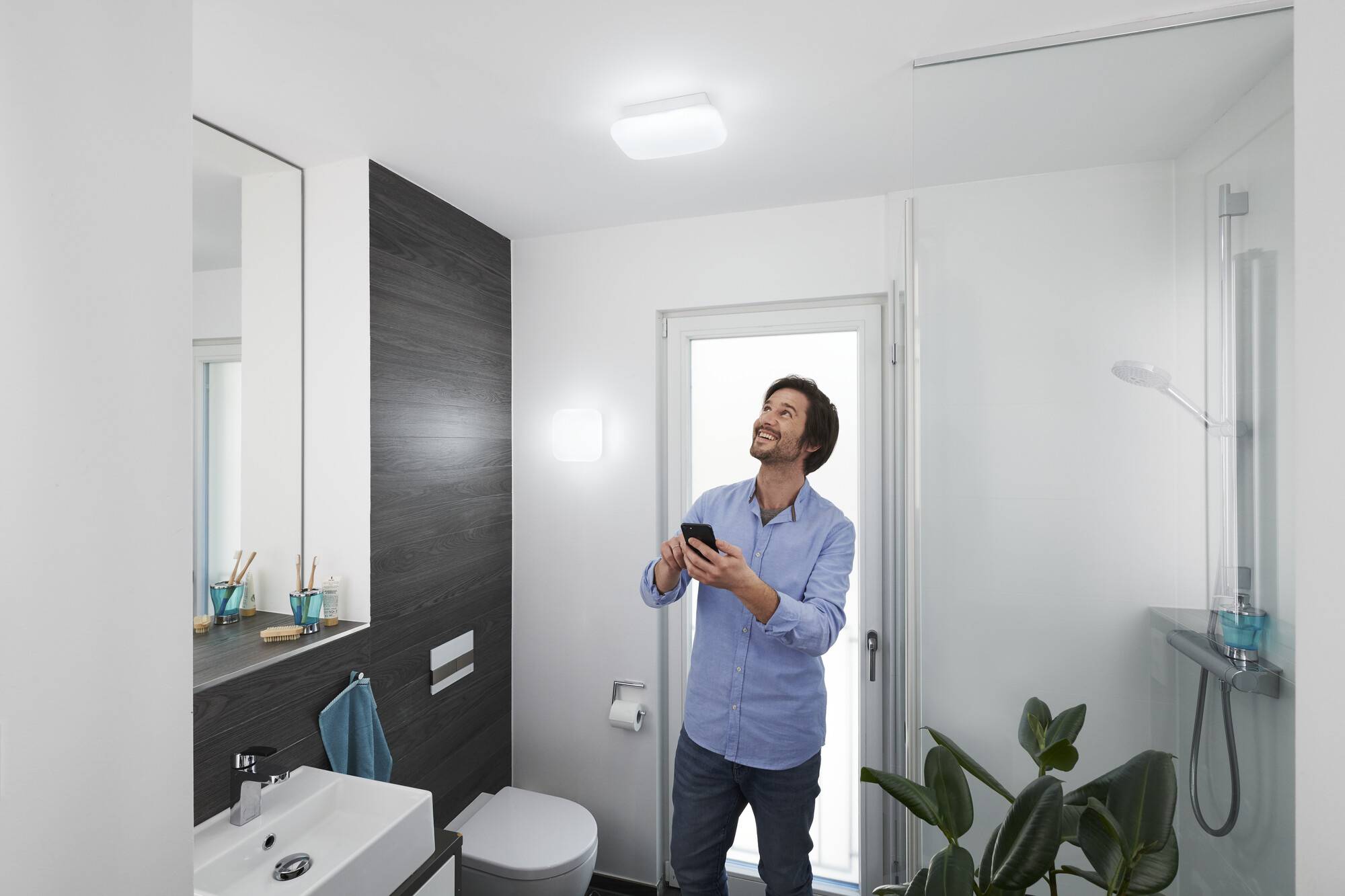 Ein Mann steht in einem modernen Badezimmer, hält ein Smartphone und schaut lächelnd zur Decke. Das Badezimmer ist hell und minimalistisch gestaltet.