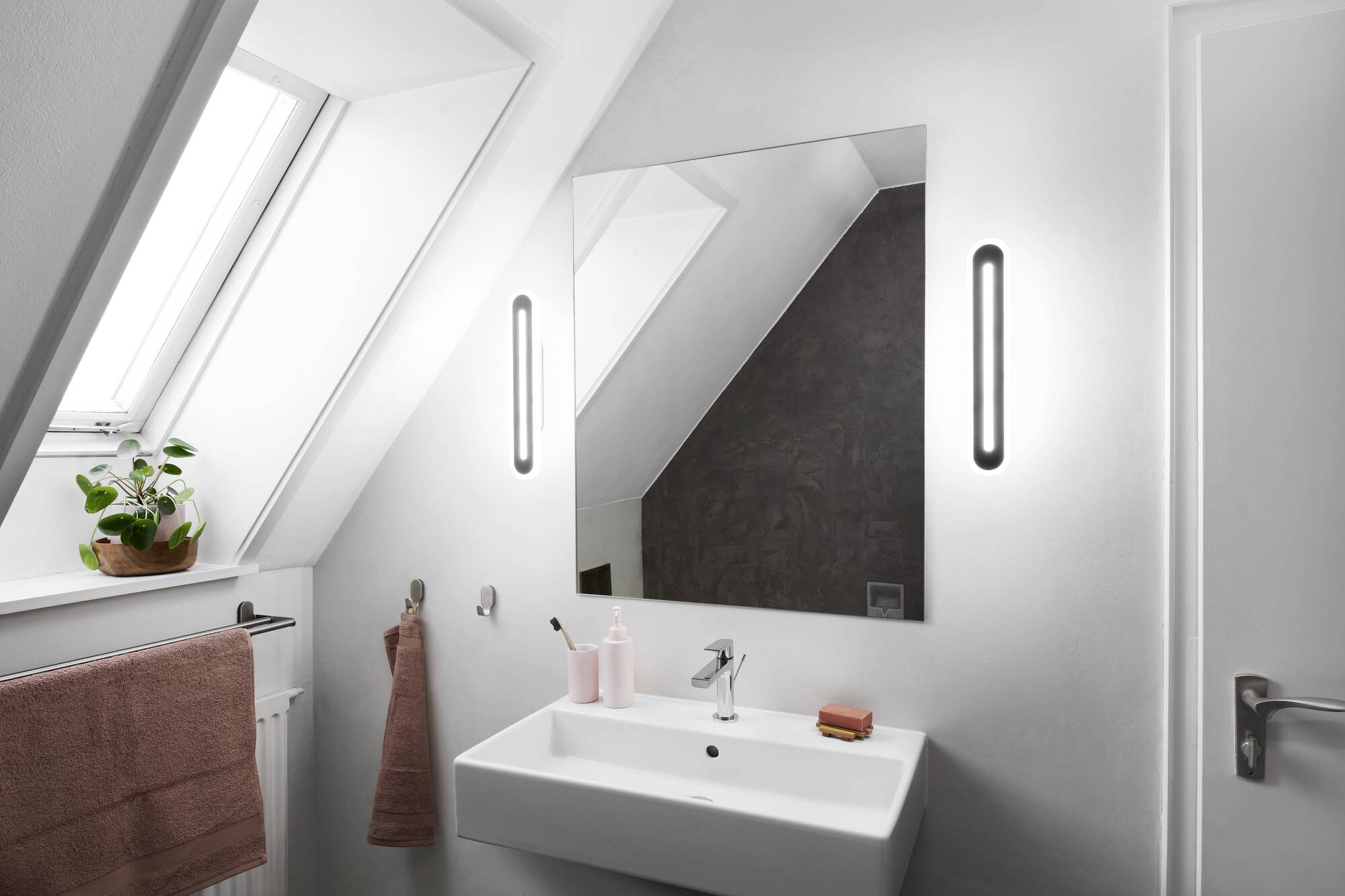 Modernes Badezimmer mit Dachfenster, großem Spiegel, Waschbecken und Handtüchern. Pflanze auf dem Fensterbrett.