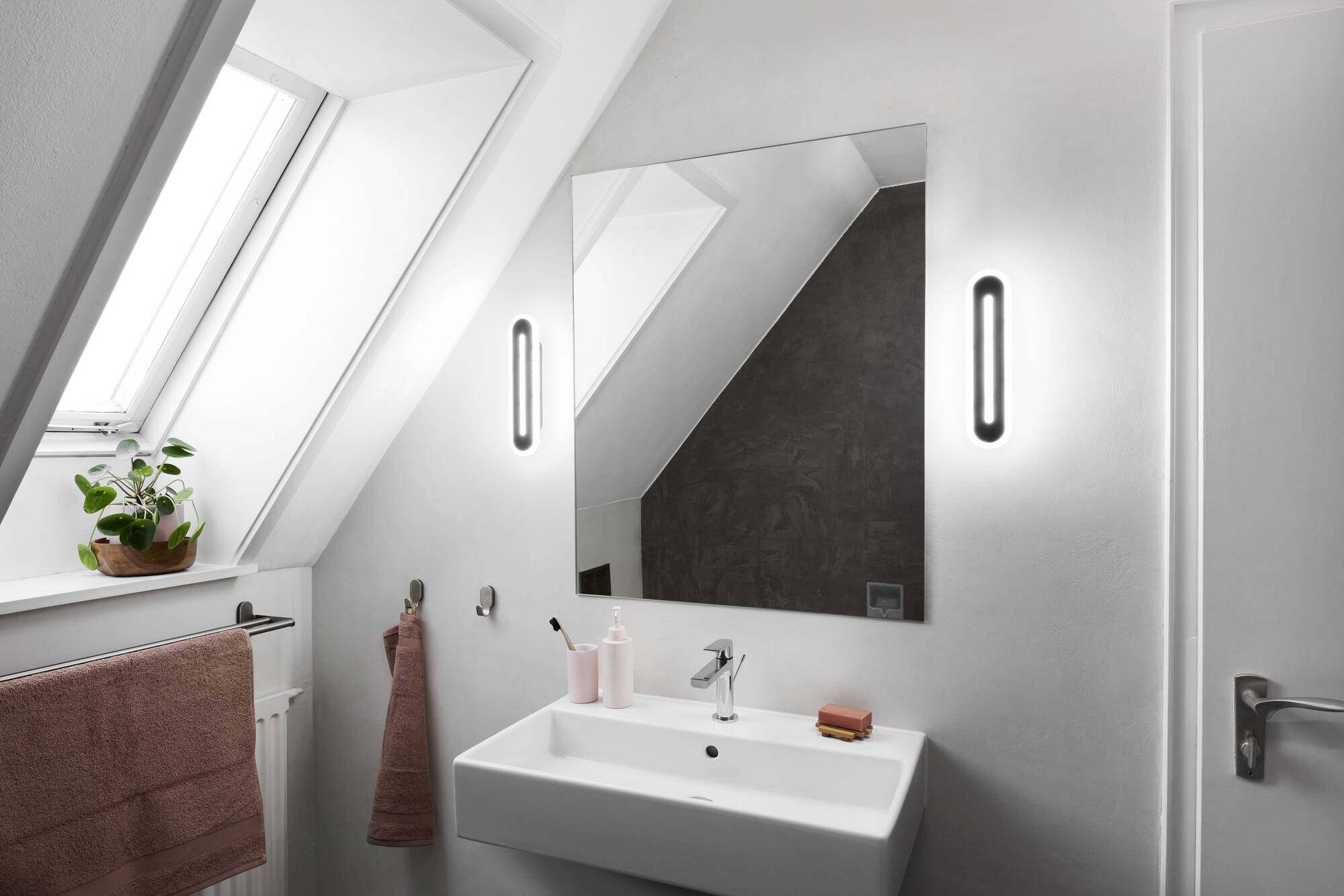 Weißes Badezimmer mit einem großen Spiegel, Waschbecken, Seifenspender, Handtuchhalter und Pflanzen am Fensterbrett unter Dachschräge.