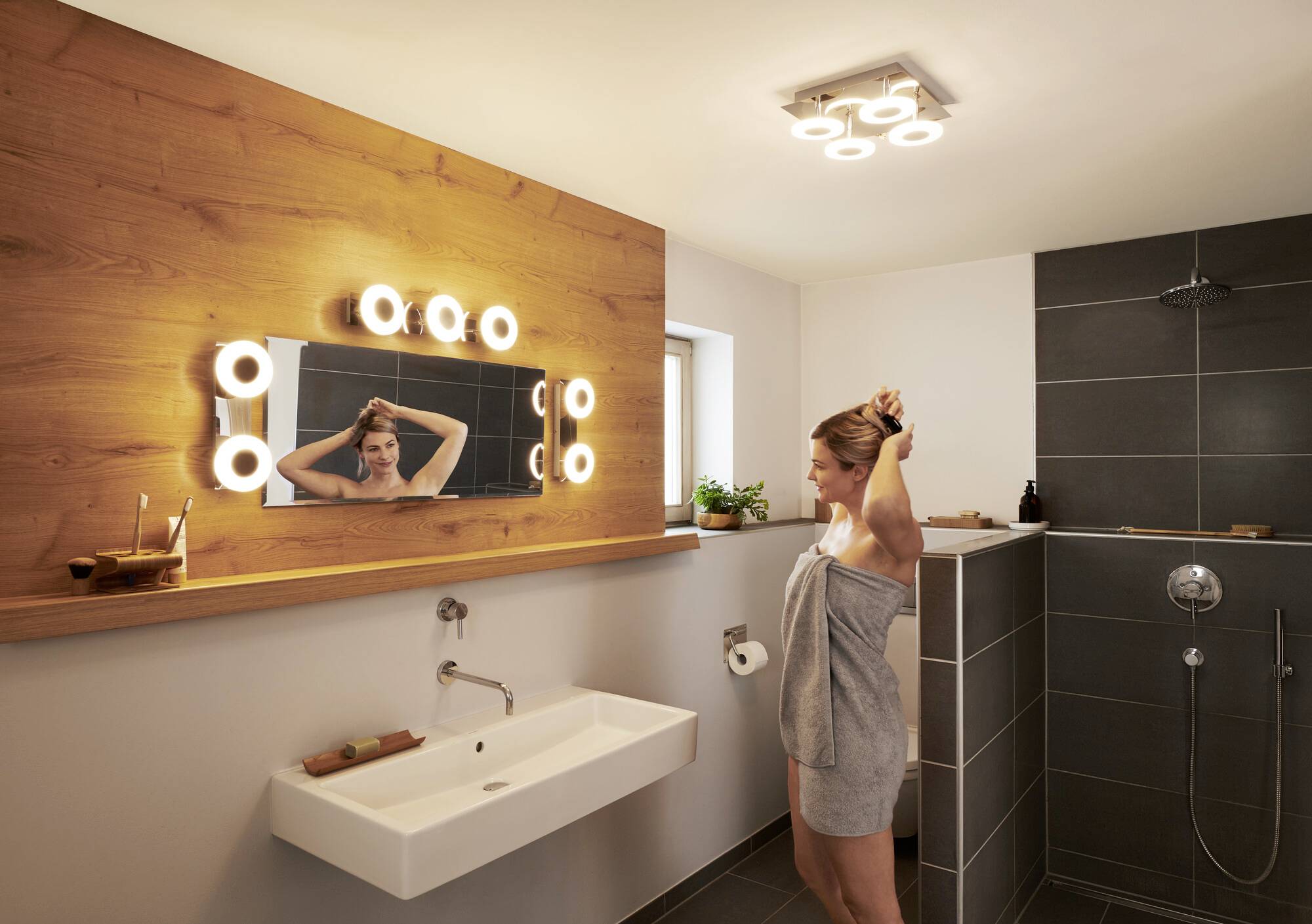Eine Frau steht in einem modernen Badezimmer vor einem beleuchteten Spiegel und bindet sich die Haare. Das Bad ist stilvoll eingerichtet.