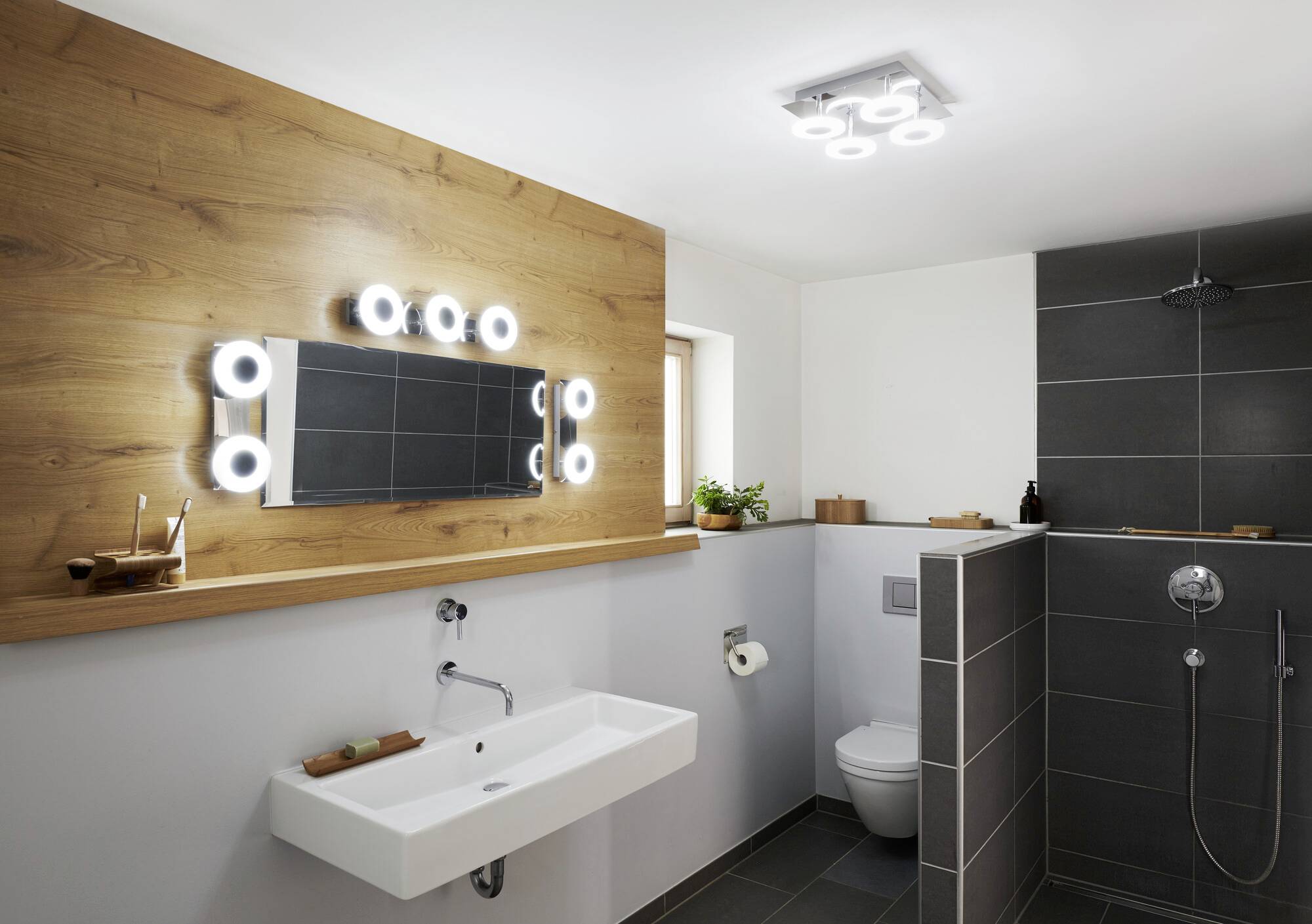 Ein modernes Badezimmer mit beleuchtetem Spiegel, Waschbecken, Toilette und begehbarer Dusche. Holz- und Grautöne dominieren.
